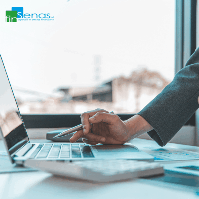 Mano con penna sopra una tastiera di laptop, accanto a documenti e calcolatrice su una scrivania.