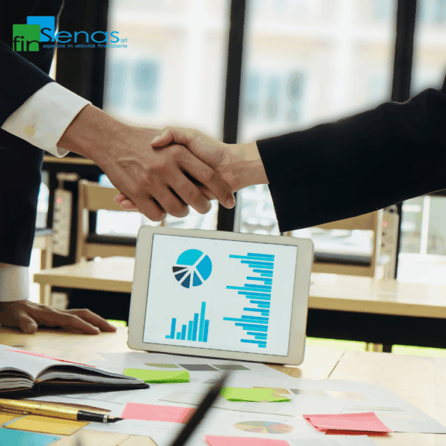 Stretta di mano tra due professionisti con grafici finanziari su tablet e documenti sul tavolo.