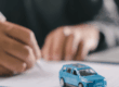 Modellino di un’automobile appoggiato su documenti mentre una persona compila una pratica, rappresentativo del credito al consumo per finanziare l’acquisto di beni e servizi.